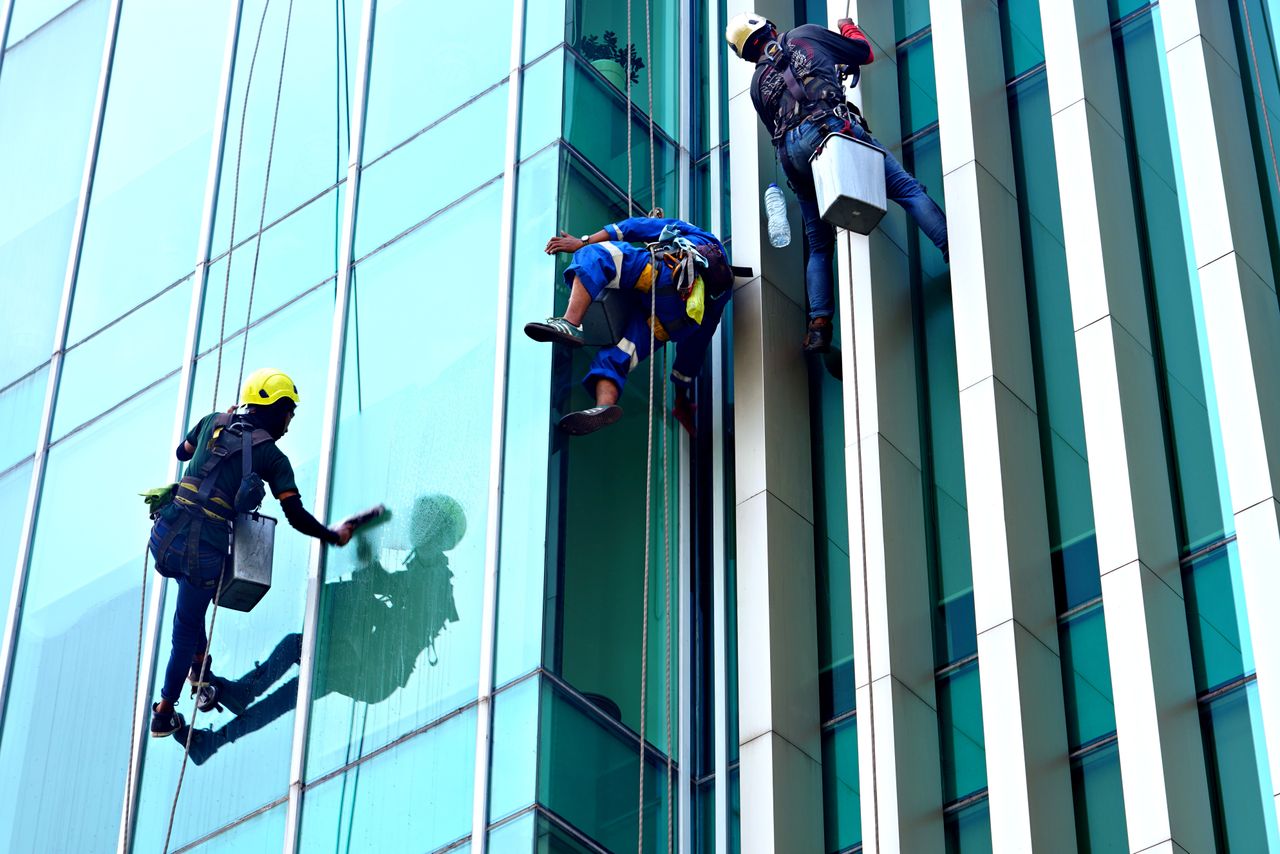 hero-img-01 Three window cleaners suspended on ropes clean a modern glass building facade during the day.